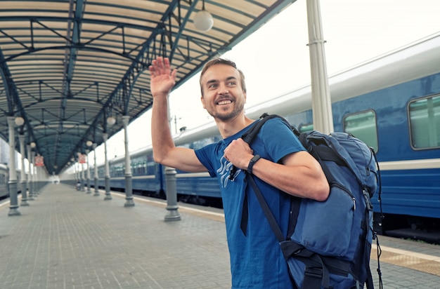 Glücklicher Manntourist mit Rucksackstand auf Bahnhofsplattform, Freunde grüßend oder verabschieden sich und bewegen seine Hand wellenartig. Mit dem Zug reisen.