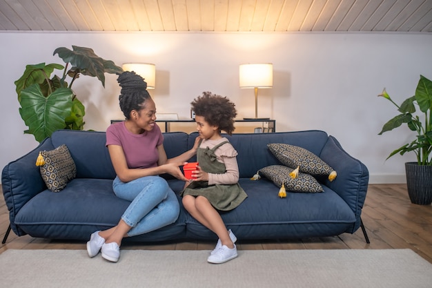 Glücklicher Moment. Kleines dunkelhäutiges lockiges süßes Mädchen, das einer glücklichen jungen Mutter, die zu Hause auf dem Sofa sitzt, ein Geschenk in einer roten Schachtel gibt