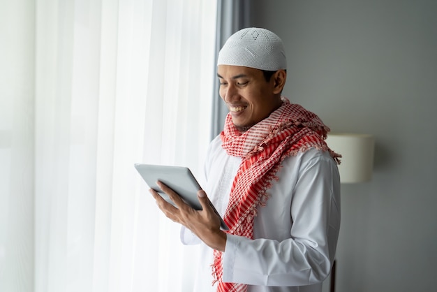 Glücklicher muslimischer Geschäftsmann, der lächelt, während Tablet-PC nahe Fenster verwendet