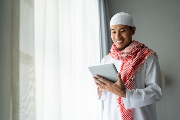 Glücklicher muslimischer Geschäftsmann, der lächelt, während Tablet-PC nahe Fenster verwendet