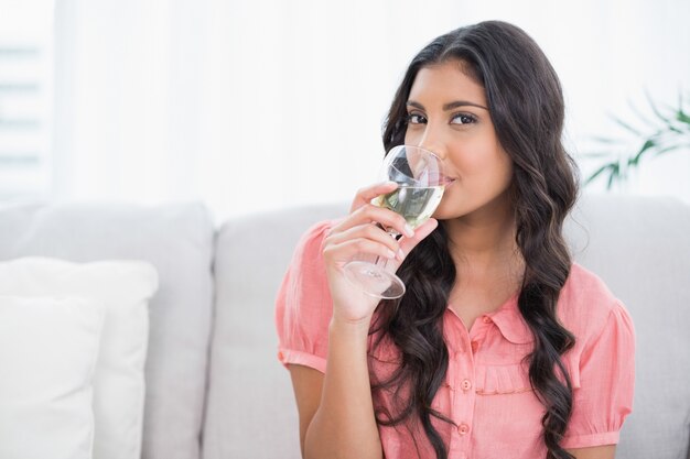 Glücklicher netter Brunette, der auf trinkendem Weißwein der Couch sitzt