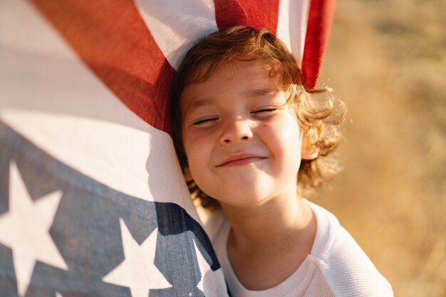 Glücklicher Patriot des kleinen Jungen, der auf dem Gebiet mit amerikanischer Flagge läuft. USA feiern den 4. Juli