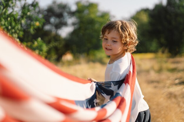 Glücklicher patriot des kleinen jungen, der auf dem gebiet mit amerikanischer flagge läuft. usa feiern den 4. juli