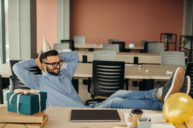 Glücklicher positiver bärtiger Büroangestellter mit Kegelhut, der mit Laptop und Geschenk mit festlichem Band am Arbeitsplatz sitzt