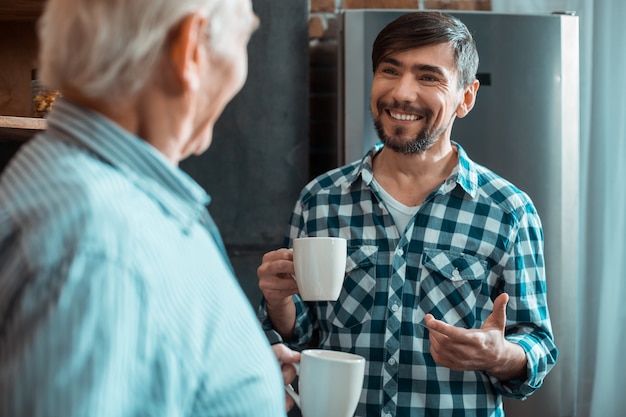 Glücklicher positiver entzückter Mann, der eine Tasse Tee hält und zu seinem Vater lächelt, während er die familiäre Atmosphäre genießt