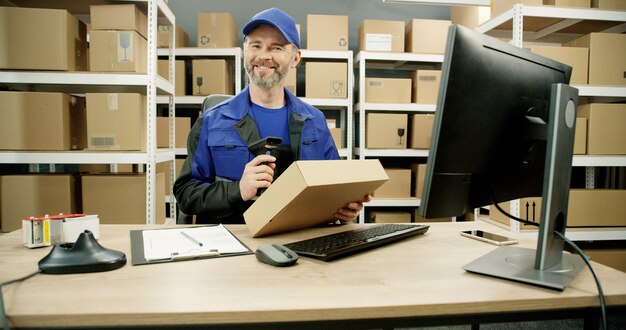Glücklicher Postbote in der Uniform, die am Computer im Postgeschäft mit Paketen arbeitet.