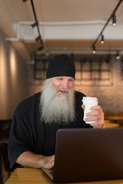 Glücklicher reifer bärtiger Mann, der Laptop benutzt und überrascht auf das Café schaut