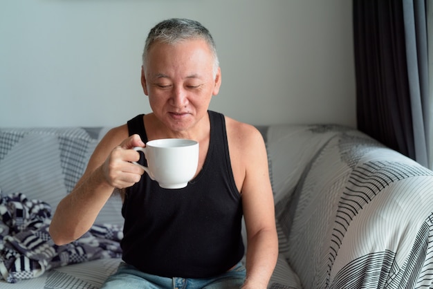 Glücklicher reifer japanischer Mann, der Kaffee im Wohnzimmer zu Hause trinkt