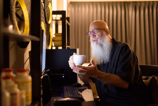 Glücklicher reifer kahler bärtiger Mann, der Kaffee trinkt, während Videoanruf bei der Arbeit von zu Hause spät in der Nacht