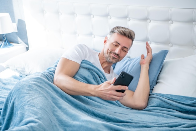 Glücklicher reifer Mann im Bett, der am Telefon chattet