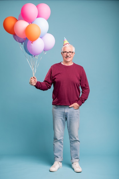 Glücklicher reifer Mann in der Geburtstagskappe und in der Freizeitkleidung, die Ballons hält, während auf der blauen Wand posierend