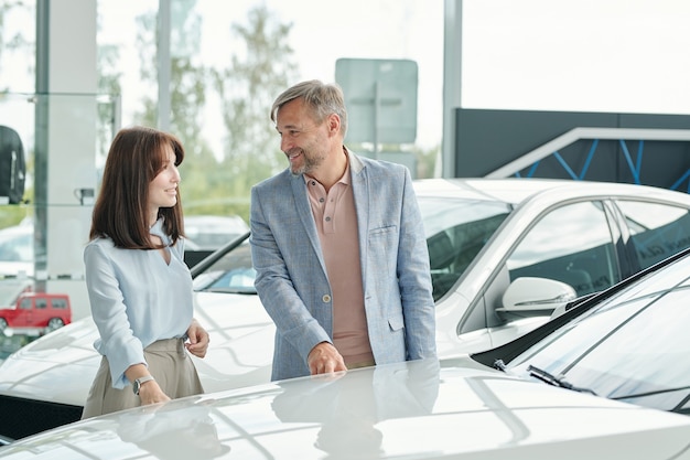 Glücklicher reifer Mann und seine hübsche elegante Frau, die ein neues Auto auswählen und sein Modell, seine Eigenschaften und seine Farbe im Autocenter besprechen