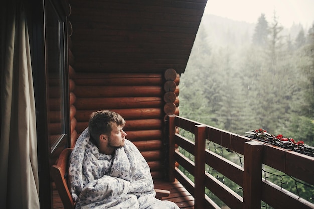 Foto glücklicher reisender, der sich in einer decke auf einer holzveranda mit blick auf wälder und berge ausruht. platz für text. hipster, der sich in einer holzhütte im wald entspannt. reisen und urlaub