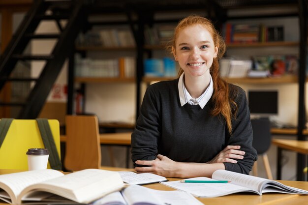 Glücklicher rothaariger Student, der am Tisch mit Büchern in der Bibliothek sitzt