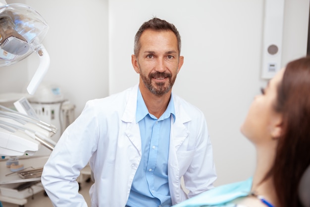 Foto glücklicher schöner reifer männlicher zahnarzt, der zur kamera lächelt, während er mit einem patienten an seiner klinik arbeitet