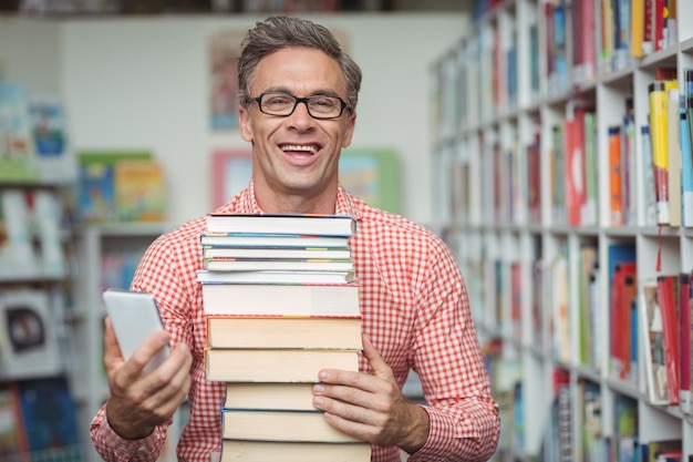 Glücklicher Schullehrer, der Stapel Bücher hält, während Handy in Bibliothek verwendet wird