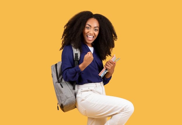 Glücklicher schwarzer Student mit lockigem Haar, der die Fäuste zusammenhält, Rucksack trägt und gelbe Kopierbücher hält