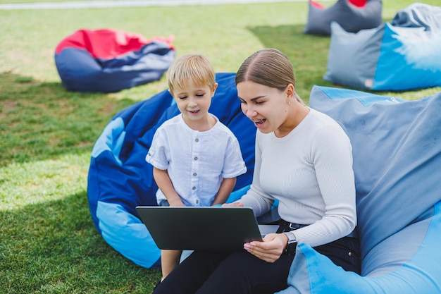 Glücklicher Sohn und Mutter sitzen in der Natur und schauen sich den Laptop an Remote-Arbeit einer jungen Mutter Familienurlaub