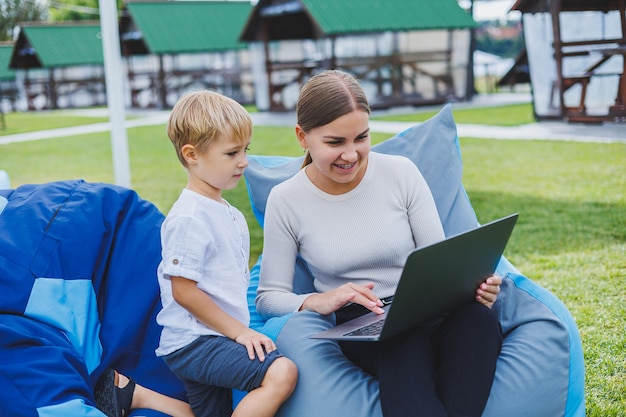 Glücklicher Sohn und Mutter sitzen in der Natur und schauen sich den Laptop an Remote-Arbeit einer jungen Mutter Familienurlaub