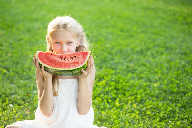 Glücklicher Sommer - schönes blondes kleines Mädchen, das Wassermelone auf einem grünen Rasen isst