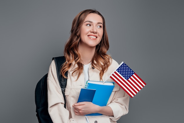 Glücklicher student, der rucksackbuch-notizbuchpass und usa-flagge hält, lokalisiert auf einer dunkelgrauen wand