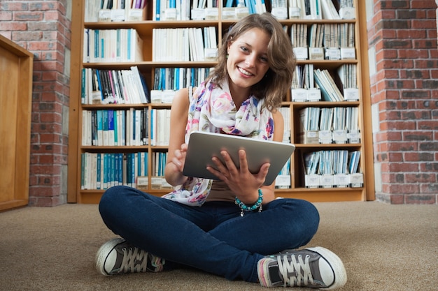 Glücklicher Student gegen Bücherregal mit Tablet-PC auf dem Bibliotheksboden