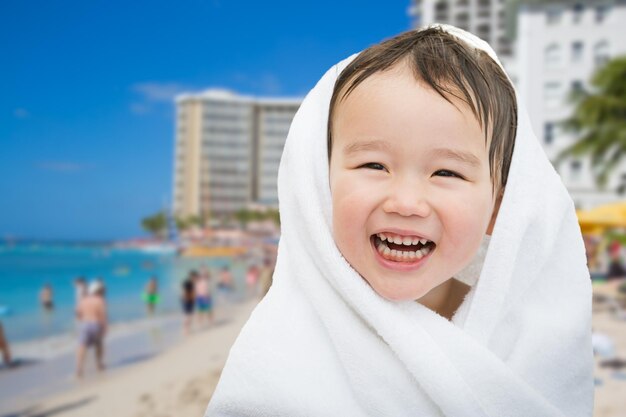 Glücklicher, süßer, gemischter Chineser und weißer Junge auf dem Strand von Waikiki, Hawaii, in ein Handtuch gewickelt.