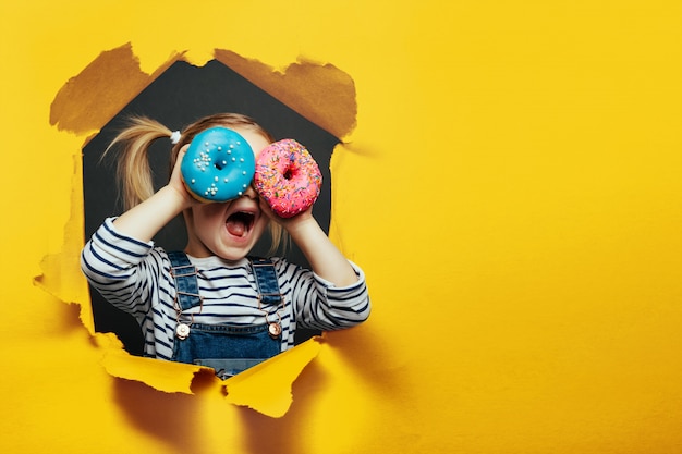 Glücklicher süßer Junge hat Spaß, der mit Donuts auf schwarzer Hintergrundwand gespielt wird.