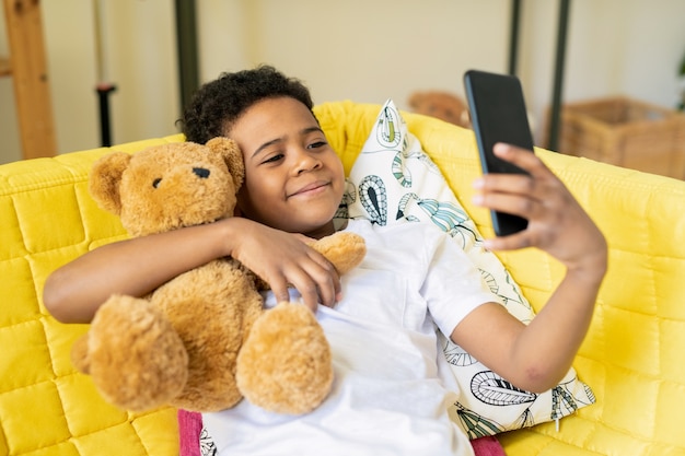 Glücklicher süßer kleiner Junge der afrikanischen ethnischen Zugehörigkeit mit Smartphone, das seinen Teddybär umarmt, während auf gelber Couch sitzen und Selfie macht