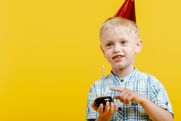 Glücklicher süßer kleiner Junge im Geburtstagshut und mit Kuchen auf Gelb.