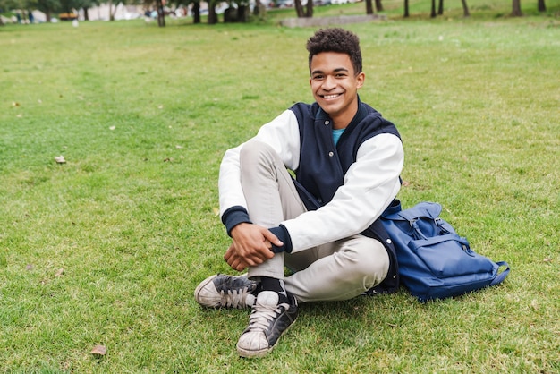 Glücklicher Teenager-Studentenjunge, der auf einem Rasen im Park sitzt