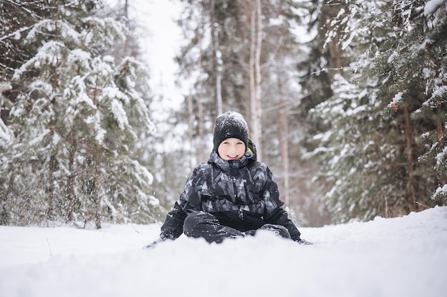 Glücklicher Teenagerjunge, der auf Schnee im Winterwald sitzt. Kind hat Spaß im Freien. Fröhlicher Jugendlicher, der bei Schneefall im Schnee spielt. Lachendes lächelndes Kind, das bei kaltem Wetter im Winterpark spazieren geht