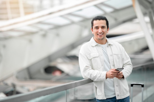 Glücklicher Tourist, der am Flughafen ein Smartphone hält, das in die Kamera lächelt