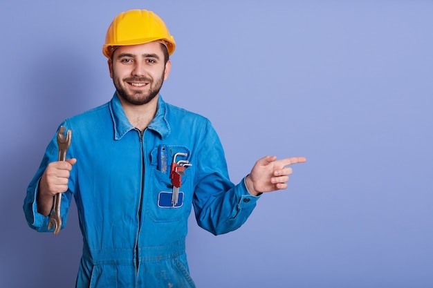 Glücklicher und lächelnder bärtiger Ingenieur mit Schlüsselwerkzeug in der Hand und mit Zeigefinger beiseite zeigend, während er dagegen steht