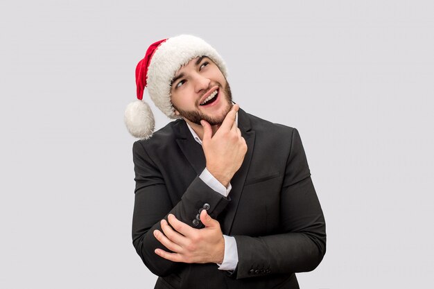Glücklicher und positiver junger Mann steht und posiert. Er sieht auf. Guy trägt Weihnachtsmütze und Anzug.