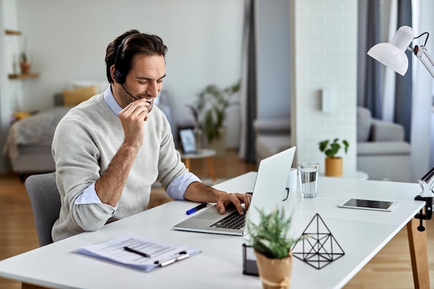 Glücklicher Unternehmer, der ein Headset trägt und mit jemandem spricht, während er zu Hause am Laptop arbeitet