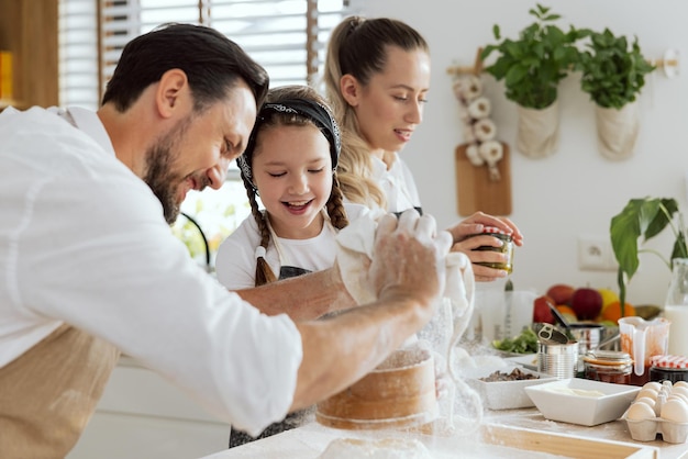 Glücklicher Vater, der Mehl aus dem Beutel auf der Holzoberfläche siebt, neugierige Tochter, die beim Beobachten des Prozesses hilft