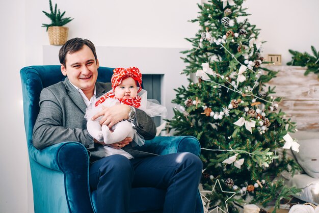 Glücklicher Vater in der eleganten Kleidung mit seiner kleinen Tochter im Kleid, das auf dem Stuhl in verziertem Feiertagsraum mit einem stilvollen Weihnachtsbaum sitzt