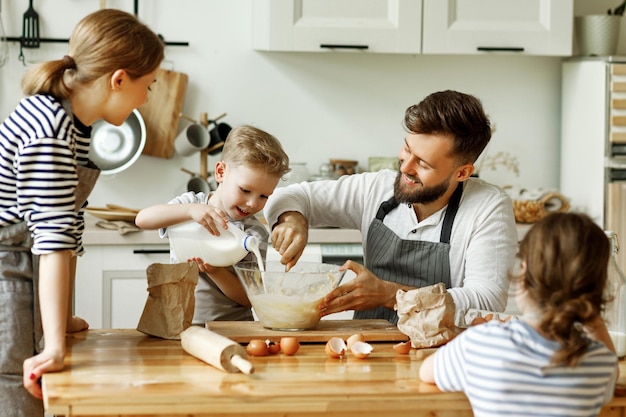 Glücklicher Vater mit kleinem Sohn, der Zutaten für Teig mischt, während Mutter mit Tochter beim gemeinsamen Kochen in der heimischen Küche in der Nähe steht