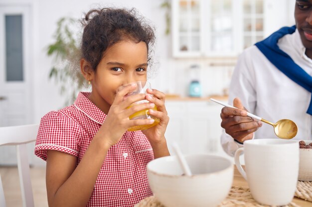 Glücklicher Vater und kleine Tochter trinken Saft zum Frühstück. Lächelnde Familie isst morgens in der Küche. Papa füttert weibliches Kind, gute Beziehung