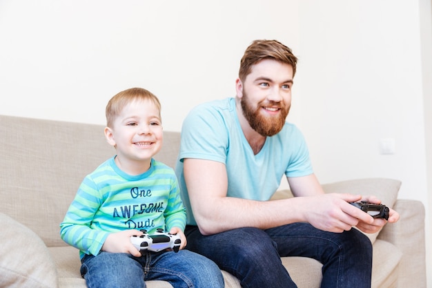 Glücklicher Vater und kleiner Sohn, die zu Hause auf dem Sofa sitzen und Videospiele spielen