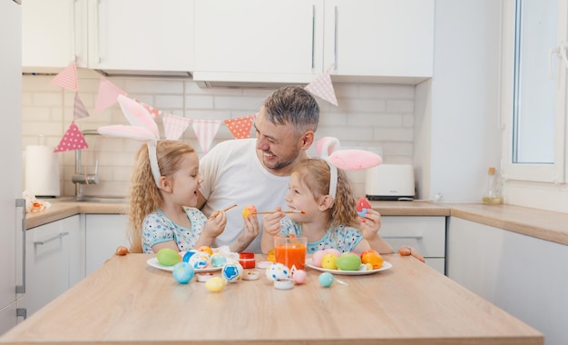 Glücklicher Vater und seine Zwillingstöchter bemalen Ostereier in der Küche und bereiten sich auf Ostern vor