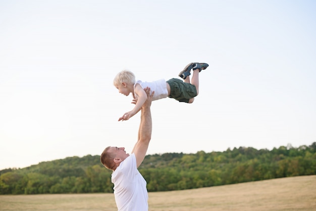 Glücklicher Vater wirft einen kleinen blonden Jungen auf einem gemähten Weizenfeld. Sonnenuntergang