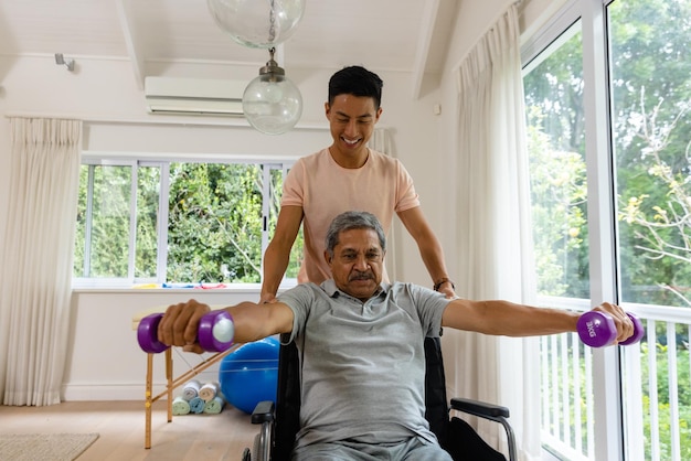 Glücklicher, vielfältiger männlicher Physiotherapeut berät und älterer männlicher Patient im Rollstuhl mit Hanteln. Physiotherapie, Gesundheitsfürsorge, Wohlbefinden und älterer Lebensstil, unverändert.