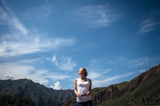 Glücklicher weiblicher Reisender in einem weißen T-Shirt gegen die schönen Berge und blauen Himmel am sonnigen Tag.