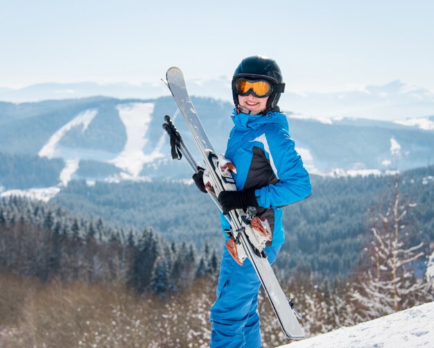 Glücklicher weiblicher Skifahrer, der zur Kamera lächelt und ihre Skier im Winterskigebiet hält