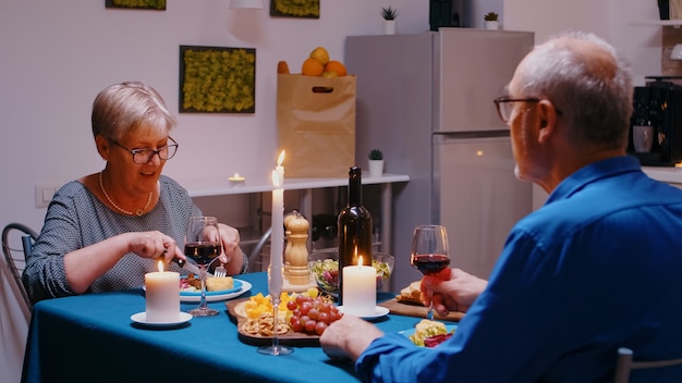 Glückliches älteres altes Ehepaar, das am Esstisch sitzt und Rotwein trinkt und spricht. Zu Hause lächelnde reife Familie mittleren Alters, die ein romantisches Abendessen und angenehme Gespräche beim gemeinsamen Essen genießt.