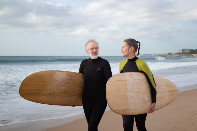 Glückliches älteres Ehepaar, das sich mit Surfbrettern ausruht. Grauhaariger Mann und Frau in Neoprenanzügen, die an der Küste entlang gehen und große Bretter halten und reden. Sportliche Aktivität und aktive Erholung für ältere Menschen