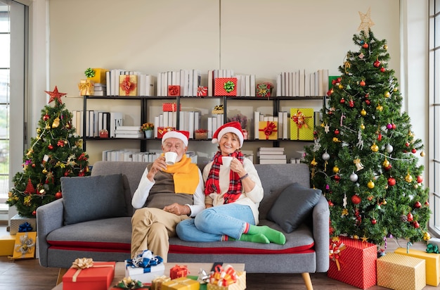 Glückliches älteres kaukasisches paar in weihnachtsmütze, das zusammen auf der sofacouch sitzt und heißes getränk zu hause mit verziertem weihnachtsbaum und geschenken im gemütlichen wohnzimmer hält. urlaub entspannen.