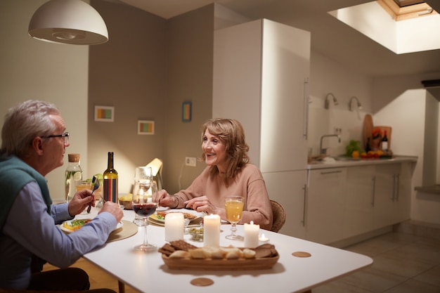 Glückliches älteres Paar, das am Tisch in der häuslichen Küche sitzt, sie haben festliches Abendessen